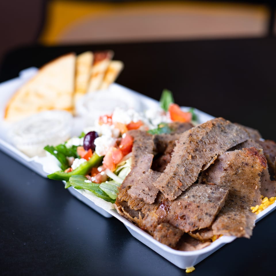 A gyro plate with sliced meat, Greek salad, pita bread, and dipping sauces in a white tray.