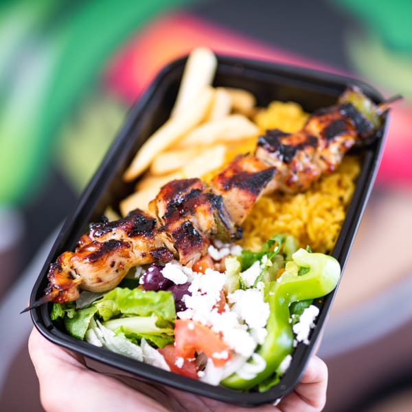Grilled chicken skewer, rice, fries, and a fresh salad with feta in a black to-go container.