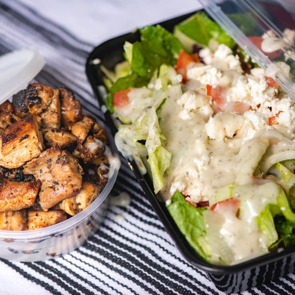Grilled chicken pieces in a clear container next to a salad with dressing and feta cheese.