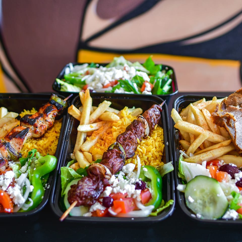 Three takeout containers filled with grilled skewers, rice, fries, and fresh salads.